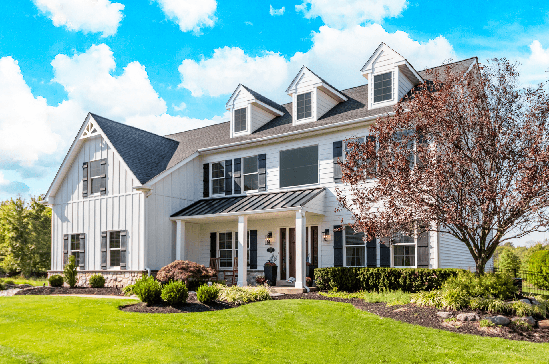Large white farmhouse-style home with board and batten siding, black shutters, covered front porch, dormer windows, manicured landscaping, and a lush green lawn beneath a bright blue sky with scattered clouds.