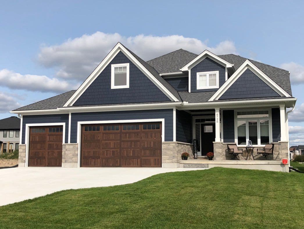 Modern two-story home with dark blue siding, white trim, stone accents, three-car garage with wood-look doors, covered front porch, and a neatly maintained front lawn under a bright blue sky.